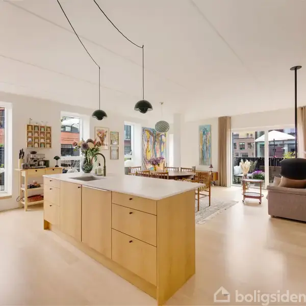Et moderne køkken-alrum med en central ø i træ, oplyst af hængelamper. I baggrunden ses en stue med sofa, kunst på væggene og store vinduer med udsigt.