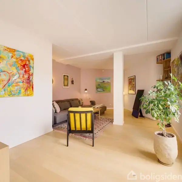 Stue med sofa og orange kunstværk på væggen, lys trægulv, grøn plante i højre hjørne, moderne indretning og flere malerier, der skaber en farverig og indbydende atmosfære.