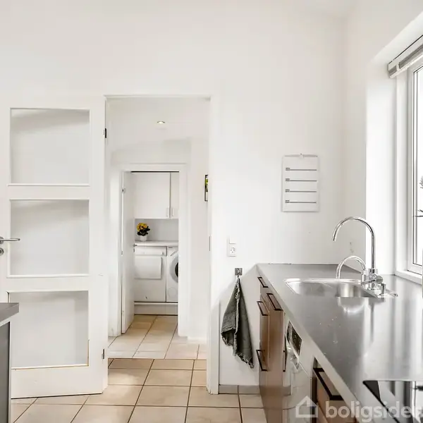 Et moderne køkken med en sort bordplade, hvor der står en vase med blomster. En håndvask er monteret under vinduet. I baggrunden ses en hvid dør og en vaskemaskine.