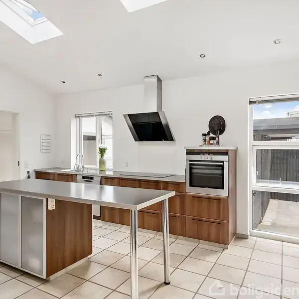 Moderne køkken med stålbord og køkkenø; ovenlysvinduer lyser rummet op. I baggrunden er der træskabe og indbygget ovn. En dør og vinduer fører ud til terrasseområdet.