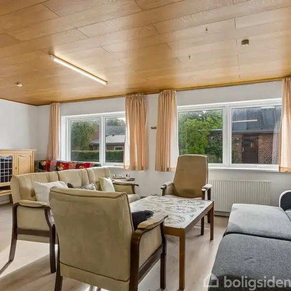 Stue med beige lænestole og sofa omkring et sofabord, placeret foran store vinduer med lyse gardiner. Træloft med enkel belysning. Skænk og maleri i hjørnet.