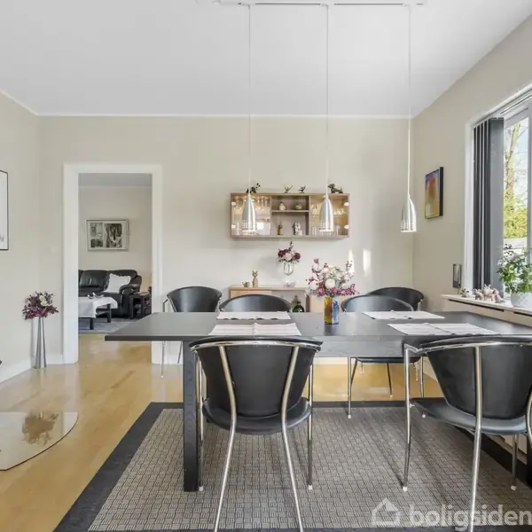 Et moderne spisebord med seks sorte stole omkring. En pejs brænder i hjørnet. Rummet har lyse vægge, trægulv og stort vindue til haven. Hylde med dekorationer bagved.