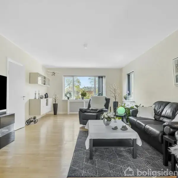 En moderne stue med sorte lædersofaer og et sofabord i centrum. Et stort fladskærms-tv hænger på væggen. Rummet har lyse trægulve og store vinduer med grøn udsigt.