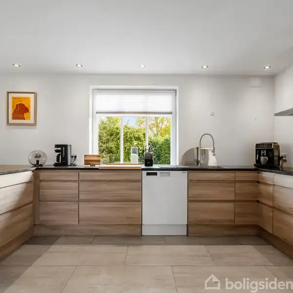 Lysebrunt køkken med sorte bordplader, udstyret med opvaskemaskine, kaffemaskine og ur. Et vindue med udsigt til træer er centralt placeret, og en plante er på venstre bordplade.