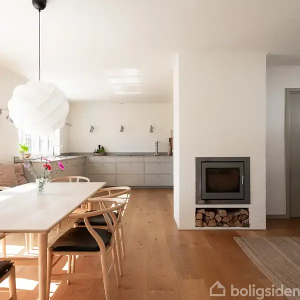 Spisebord med stole står i et lyst rum ved siden af et moderne køkken. Der er en pejs med brænde under, og bokhylder på væggen.