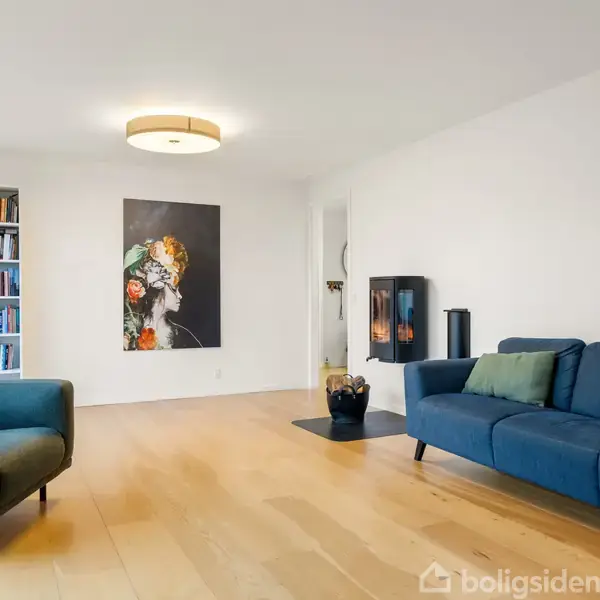En stue med en blå sofa og en lænestol, begge med puder, står på et trægulv. Til venstre ses en bogreol, og en moderne pejs findes i baggrunden. En stor blomstermaleri hænger på væggen.