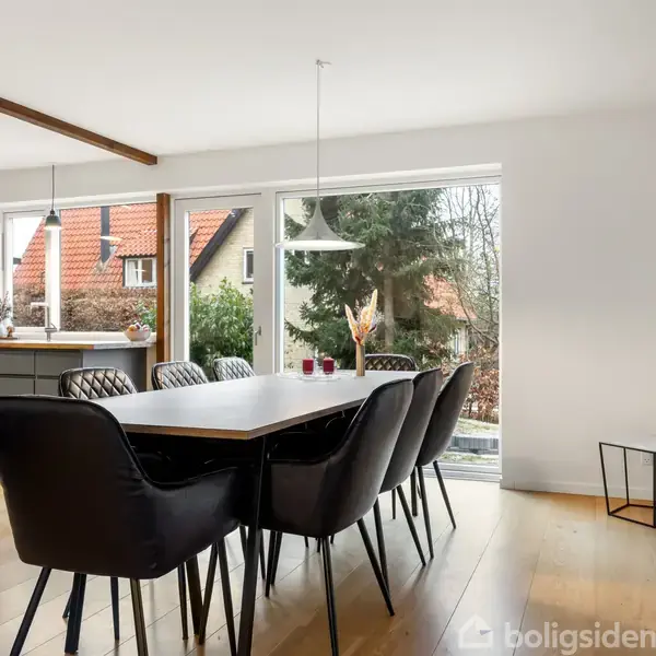 Et langbord med otte sorte stole står i en spisestue. Store vinduer giver udsigt til en have med grønne træer. En minimalistisk indretning præger rummet.
