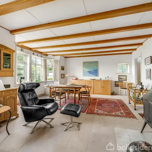 Stue med lænestol og skammel, placeret ved vinduer. Reol står mod væggen. Spisebord og tæppe ses centralt. Pejs til højre skaber varme i rummeligt opholdsområde.