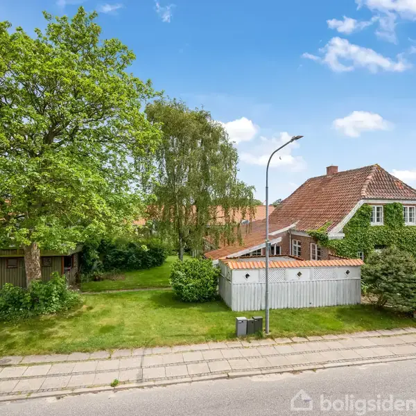 Hus med rødt tag er omkranset af grøn beplantning, herunder træer og buske. Ved siden af ses en garage med bil. Det hele ligger i et fredeligt boligområde på en solrig dag.