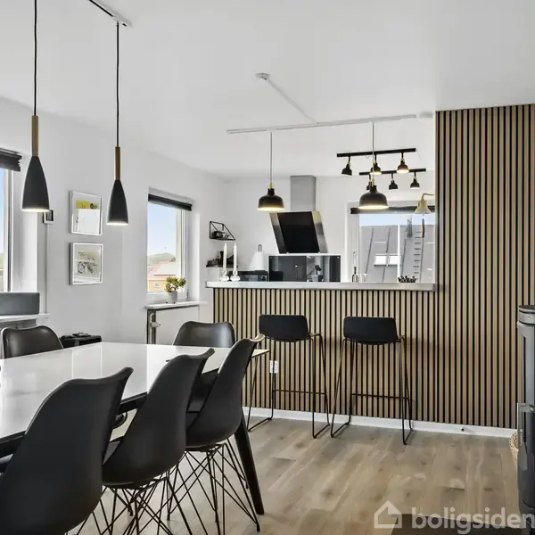 En moderne spisestue med et sort spisebord og stole, belyst af hængelamper. Åben forbindelse til køkkenet med barstole og en stribet væg. Vinduer lukker naturligt lys ind.