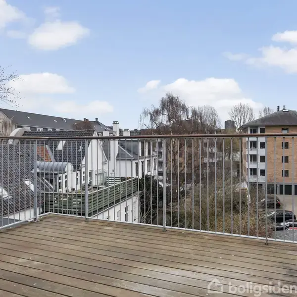 En altan med trægulv og metalrækværk står stille. Den omkranses af bybebyggelse med både moderne og ældre bygninger under en blå himmel med lette skyer.