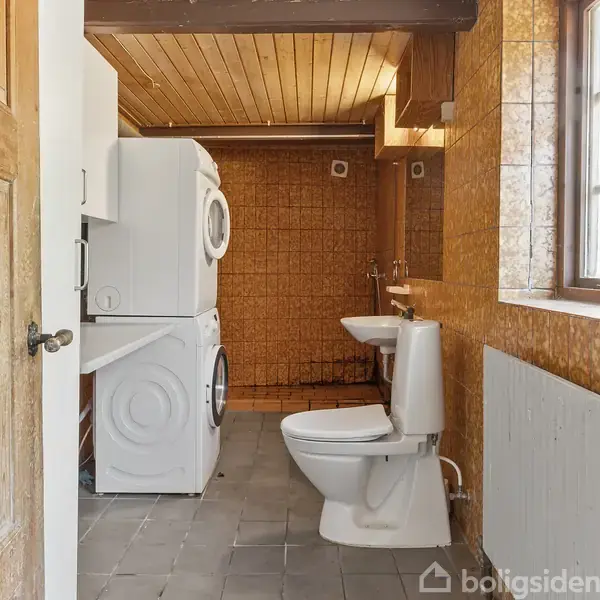 En vaskemaskine og tørretumbler er stablet i et badeværelse med brune fliser. Til højre er et toilet ved siden af et håndvask. Lyset kommer ind fra et vindue.