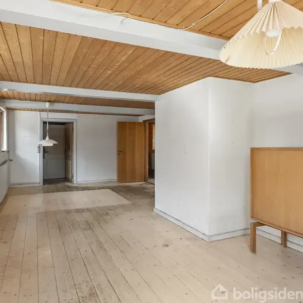 Et tomt rum med trægulv og træloft. En hvid radiator og flere vinduer ses til venstre. Der hænger to hvide lamper fra loftet, og et skab står til højre.