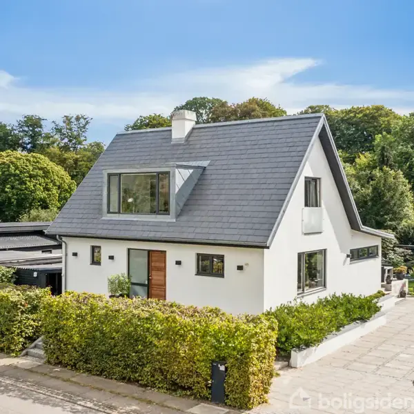 Hvidt hus med sort tag står stille ved siden af en bil på en garageindkørsel omgivet af grønne buske og frodig skov i baggrunden.