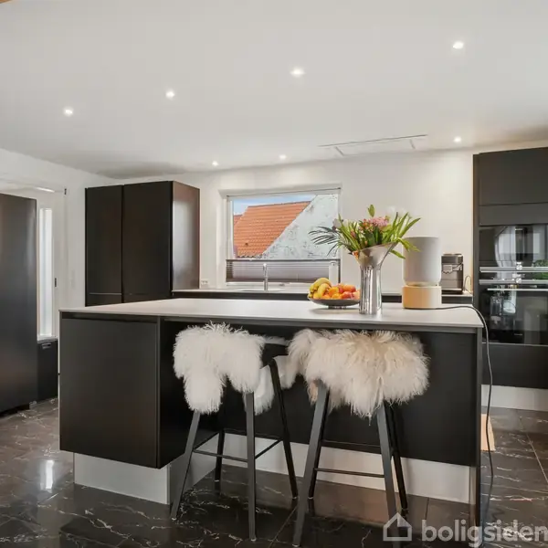 Et moderne køkken med en stor, sort ø i midten, dekoreret med blomster og skamler dækket af pels. Køleskab til venstre og indbyggede ovne til højre.