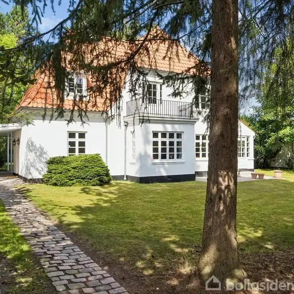 Et hvidt hus med rødt tegltag står i en have omringet af træer. En bil er parkeret under en carport til venstre. Græset er grønt og velplejet.