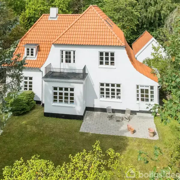 Hvidt to-etagers hus med rødt tegltag står i grønt haveområde. En terrasse med borde og stole er foran huset, omgivet af tætliggende træer og buske.