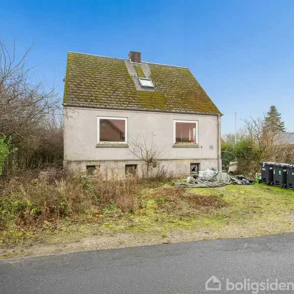Et ældre, forladt hus med mosdækket tag står tomt i en vildtvoksende have. Tre sorte affaldscontainere er placeret foran, og der er en vej i forgrunden.