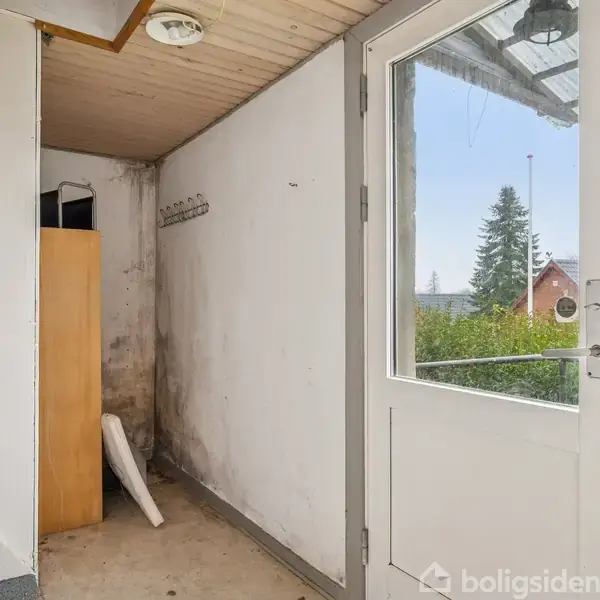 En indgang med trappe på venstre side, en trædør med vindue til højre, og et skab i hjørnet. Udenfor ses træer og et hus i baggrunden.