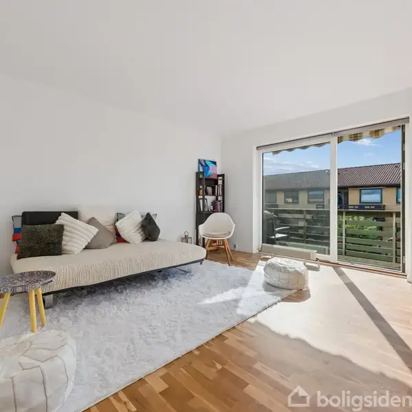 Sofa med puder står i en lys stue med trægulv. En gulvlampe og et lille bord er ved siden af, mens en kommode er ved glasskydedør ud til balkon. Ingen tekst.