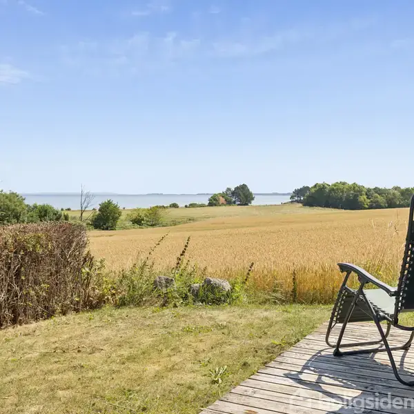 En liggestol står på en træterrasse og vender ud mod et kornmark, med hav og blå himmel i baggrunden. Grønt buskads og træer omgiver området.