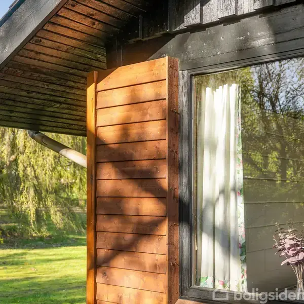 Et træhus med en udsmykket skodde ved vinduet befinder sig i en solrig have. Skarpe skygger falder på husets væg, omgivet af frodig vegetation med træer i baggrunden.