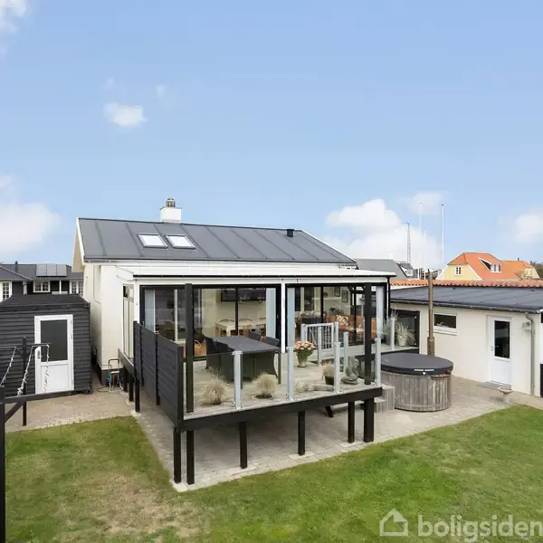 Et moderne hus med en tilbygget glasveranda. Husets værfter er omgivet af en velplejet græsplæne. Nabohuse med farverige tage ses i baggrunden under en klar, blå himmel.