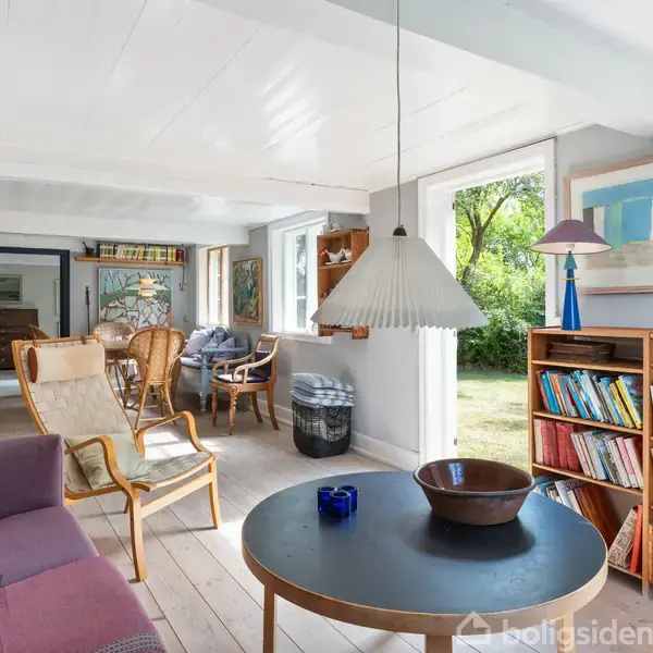 En stue med en lilla sofa og rundt bord, rummer også en reol fyldt med bøger, malerier på væggen, og store vinduer, der giver udsigt til en grøn have.