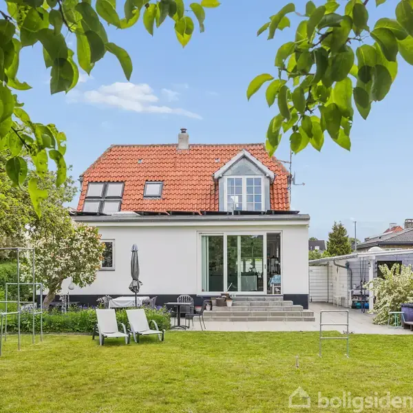 Hus med rødt tegltag og to vinduer på første sal, omgivet af grøn plæne med havemøbler. Have har træer og buske, og en terrasse er synlig nær huset.