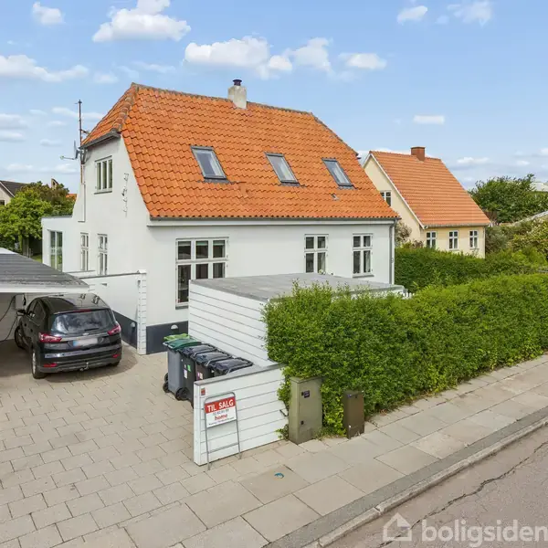 Et hvidt, to-etagers hus med rødt tegltag står ved en vej. En bil er parkeret under en carport ved siden af huset. Grønt levende hegn omgiver ejendommen. Skilt ved indkørslen: "Til salg" med kontaktinformationer fra en ejendomsmægler.