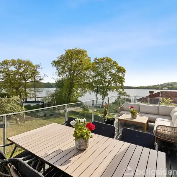 Terrasse med træbord og stole; blomster i krukker på bordet; udsigt over sø omgivet af træer og huse; klar blå himmel.