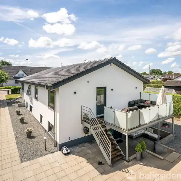 Hvid villa med sort tag og glasbalkon. En trappe fører op til terrassen. Omkringliggende grusvej og hække i et fredeligt boligkvarter med let skydække på himlen.