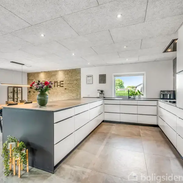 Lyst køkken med hvide skabe og en sort køkkenø. En vase med blomster står på bordet. På væggen ses ordet "HOME". Store vinduer giver udsigt til en grøn have.