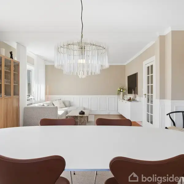Ovalt spisebord med brune stole står i en lys stue. En elegant lysekrone hænger over bordet. Stuen har sofa, vitrineskab, fladskærms-TV og store vinduer med hvide gardiner.