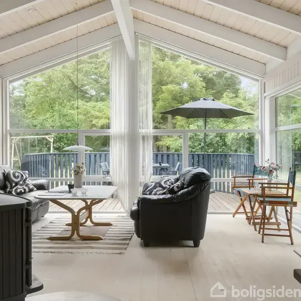 Træbeklædt stue med sorte lædersofaer og et rundt træbord, der vender mod store vinduer. Udenfor ses en åben terrasse med havemøbler og grønne omgivelser.