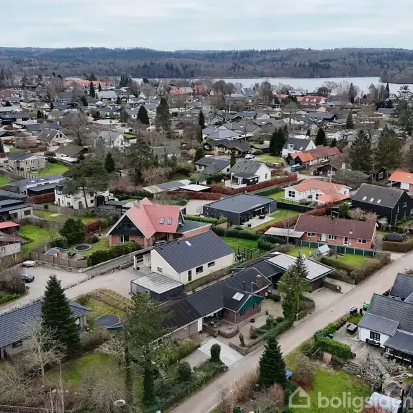 Huse ligger spredt i et villakvarter. Nogle hustage er røde eller sorte. Området er omgivet af skovklædte bakker og en sø i baggrunden under en grå himmel.