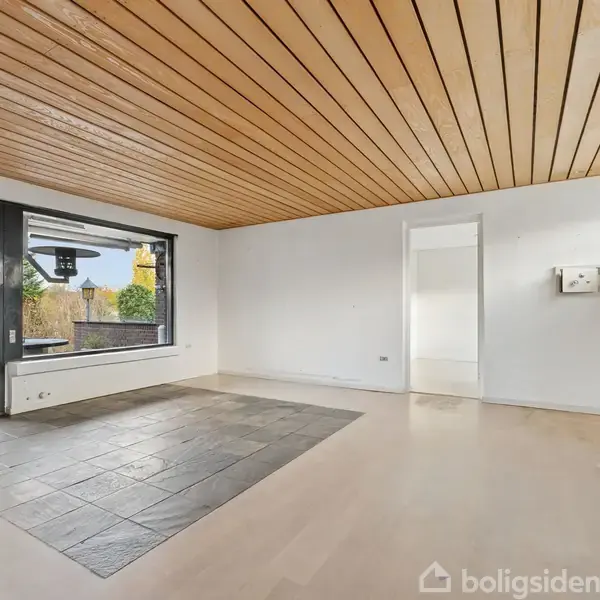 Stue med trægulv og en stor flisebelagt sektion. Et stort vindue og en glasskydedør åbner mod en terrasse og have. Lyse vægge og træloft i rum med enkel indretning.