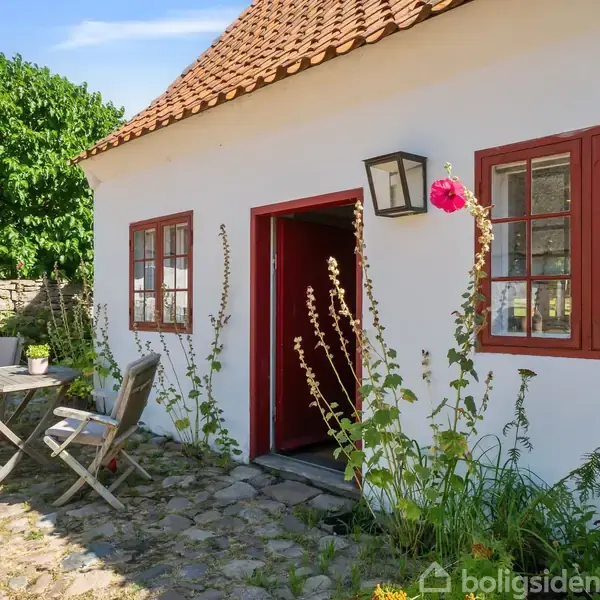 Et hvidt hus med røde vinduesrammer har en åben dør omgivet af stokroser. Foran står to stole og et bord på en brostensbelagt terrasse med grønne planter.