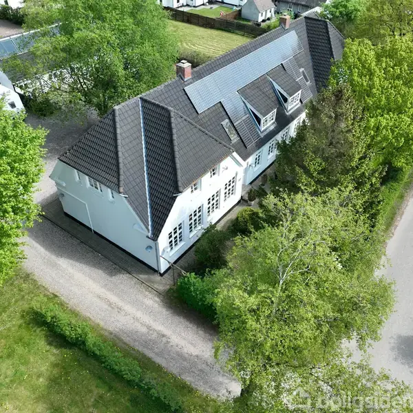 Et stort hvidt hus med mørkt tag ligger omgivet af træer. Vejen løber langs huset, mens en grussti fører op til indgangen.