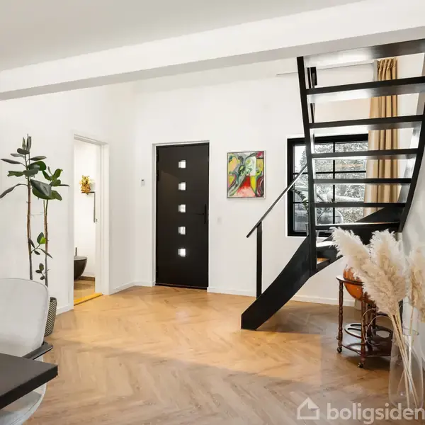 Trappe fører opad i moderne interiør med træparket, hvid væg og malerier. Dør til venstre, store vinduer til højre og planter samt pampasgræs i rummets hjørne.