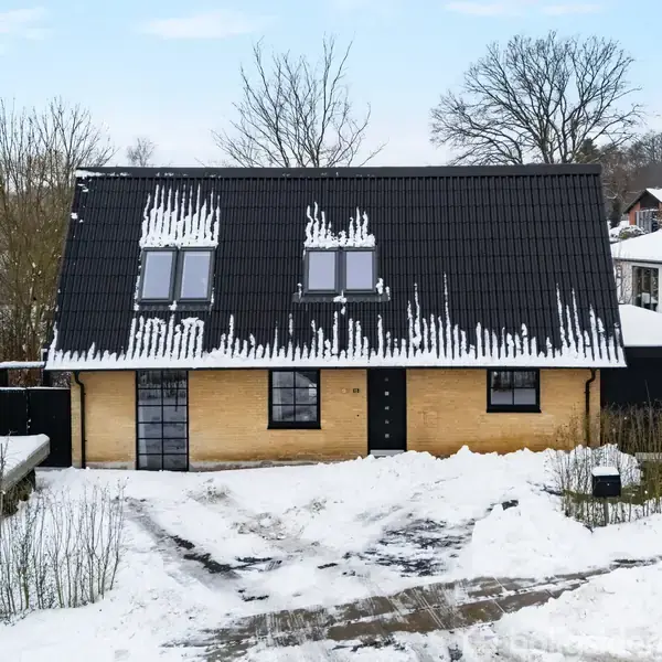 Et to-etagers hus med sort tag og gul murstensfacade står omgivet af sne. Vinduer er isplettede, og vejen er snedækket med spor af køretøjer. Træer og buske omgrænser området.