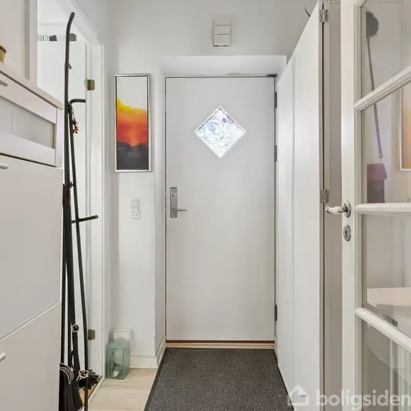 Dør står lukket i en lys gang med hvide vægge, dekorativ kunst på højre side og skoskabe til venstre på gulvtæppe og lyst gulv.