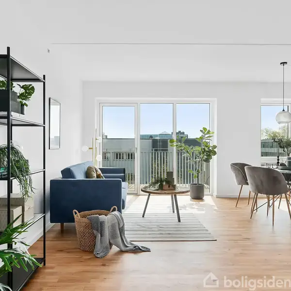 Sort reol med planter og bøger står i en moderne stue, hvor en blå sofa vender mod et sofabord. Store vinduer lukker lys ind og afslører udsigt til en altan. Ingen tekst.