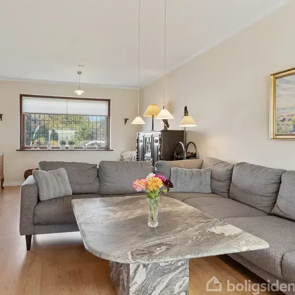 Grå sofa står i stue med et marmorbord foran. En vase med blomster er på bordet. En lampe hænger over bordet, og der hænger et landskabsmaleri på væggen.