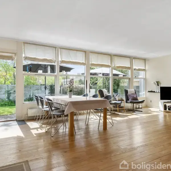 Stue med spisebord i midten, omgivet af stole, foran store vinduer med udsigt til en have. Hjørne med sofa, stort TV og gulvlampe. Sollys oplyser rummet.