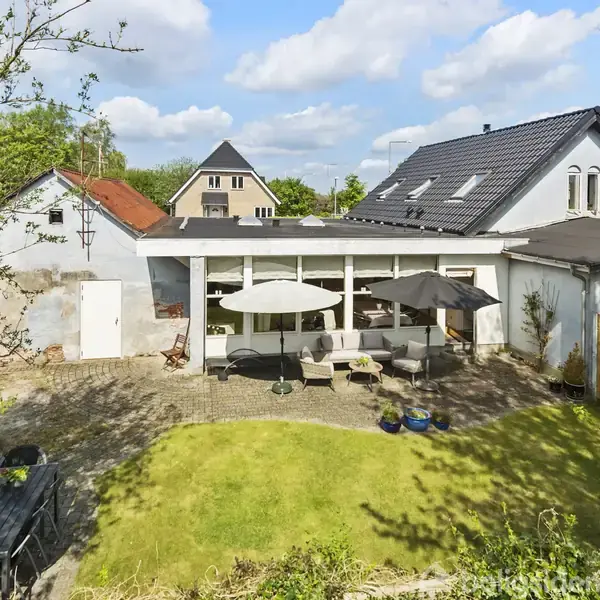 Hus med terrasse, hvor havemøbler og parasoller står på flisebelagt område. Græsplæne og grønne buske omkranser terrassen. Blåt tag med store vinduer i baggrunden under en blu himmel.
