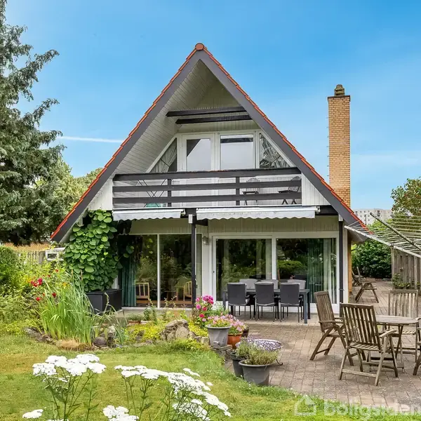 A-formet hus med balkon omkranset af en grøn have. Foran ses en terrasse med udendørs møbler. I baggrunden står træer og buske under en blå himmel.
