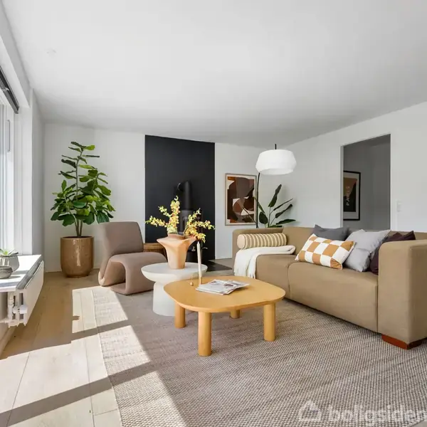 Stue med beige sofa, sofabord og lænestole. Stor plante og kunst på væggen skaber en moderne atmosfære. Store vinduer giver udsigt til en grøn have. Ingen tekst i billedet.