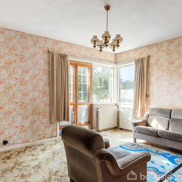 Stue med brun sofa og lænestol på et blomstret gulvtæppe. Store vinduer giver lys ind i rummet, der har blomstret tapet og gardiner. En lysekrone hænger ned fra loftet.