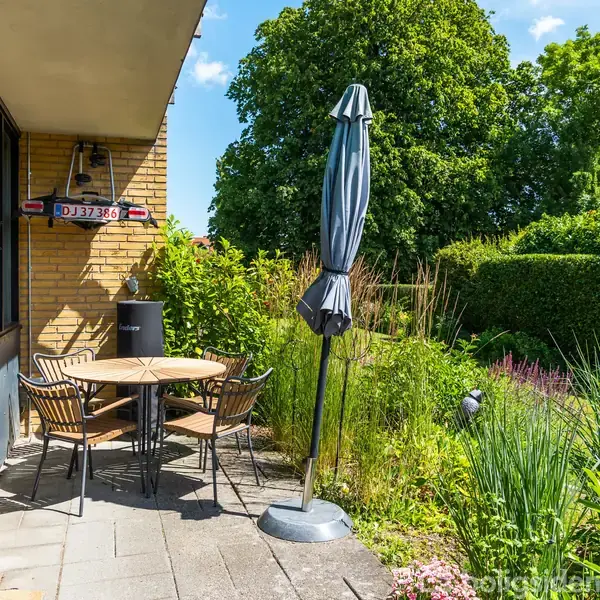 En rund træbord omgivet af fire stole står på en flisebelagt terrasse. En sammenklappet parasol er placeret ved siden af, omringet af frodig grøn vegetation og buske. En nummerplade, DJ37386, ses på væggen.
